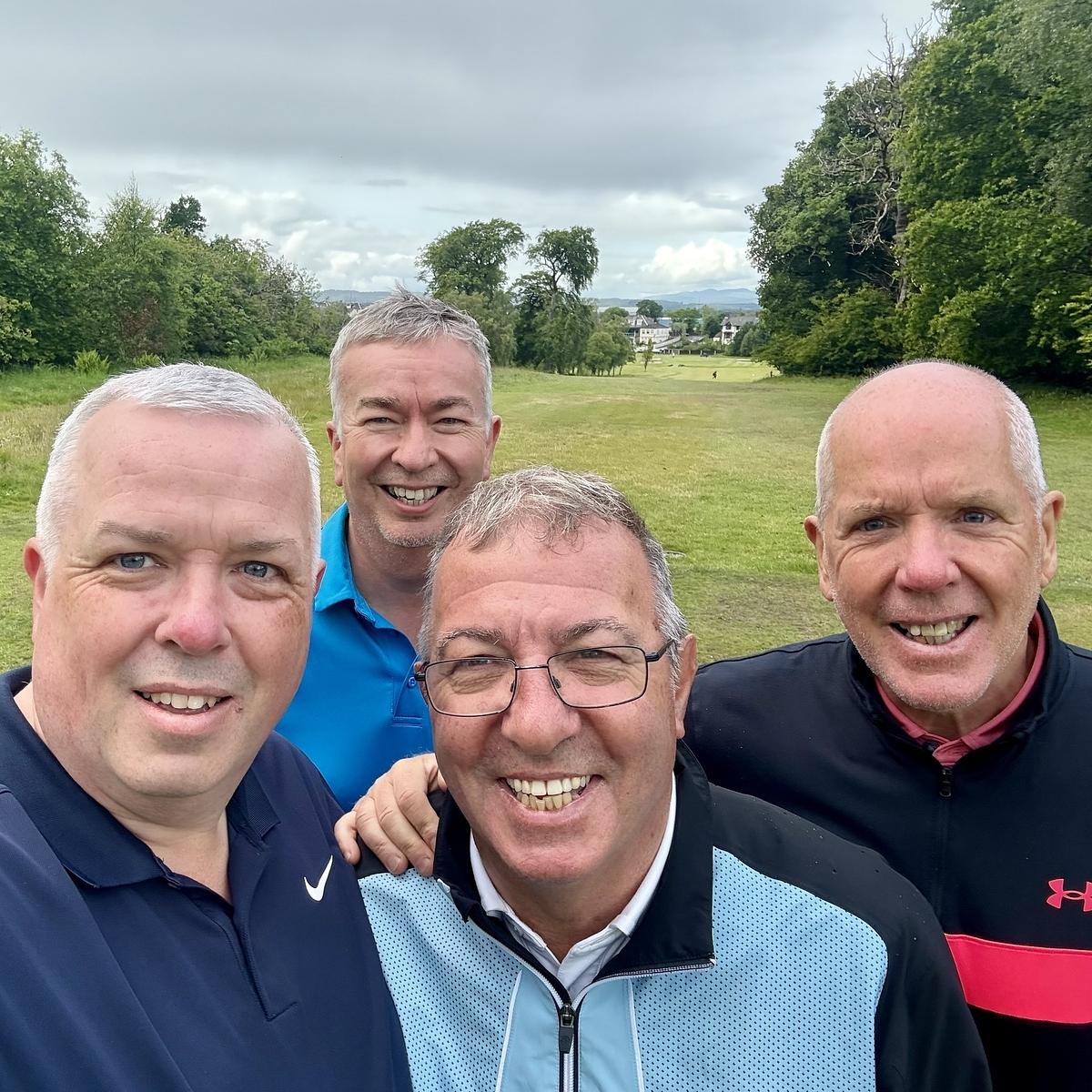 Four smiling men are posing together outdoors on a golf course with trees and a cloudy sky in the background.