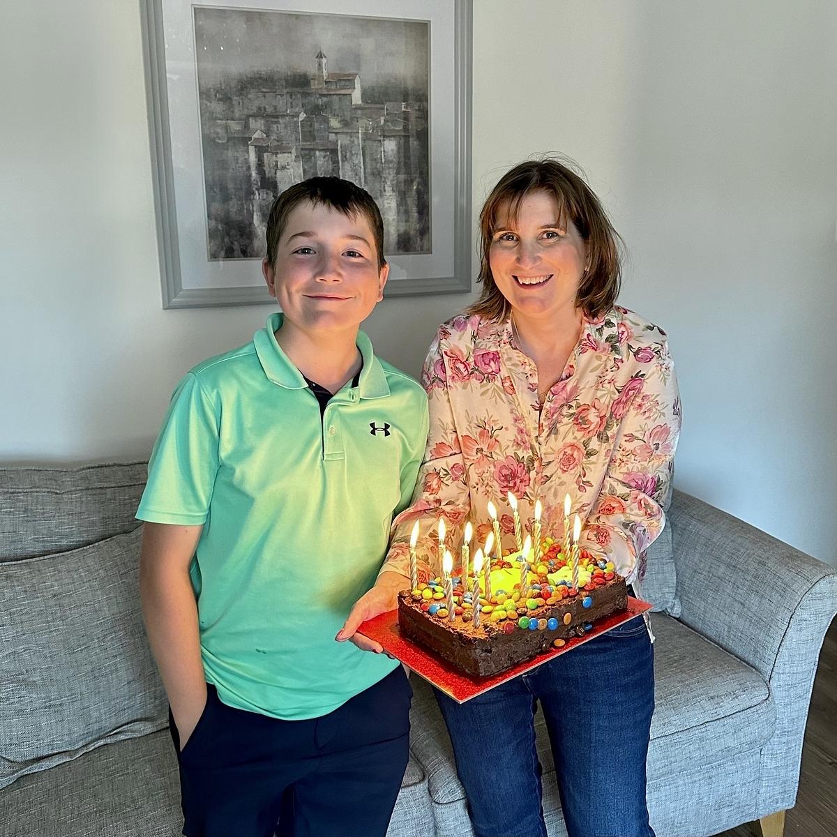 A woman and a young boy are celebrating with a lit birthday cake adorned with colorful decorations on a red tray, standing in front of a gray couch and framed artwork.