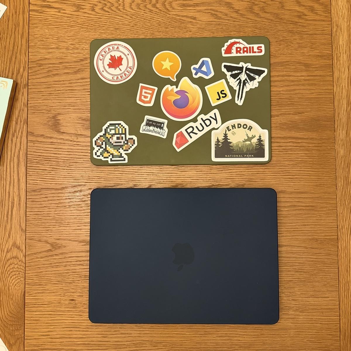 A green laptop cover with various tech-themed stickers is placed on a wooden surface, next to a closed blue laptop.