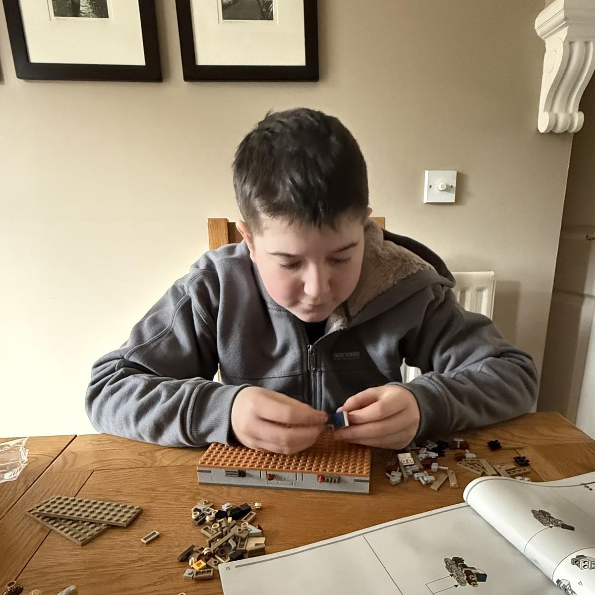 A child is assembling a Lego set at a table in a room with framed pictures on the wall.