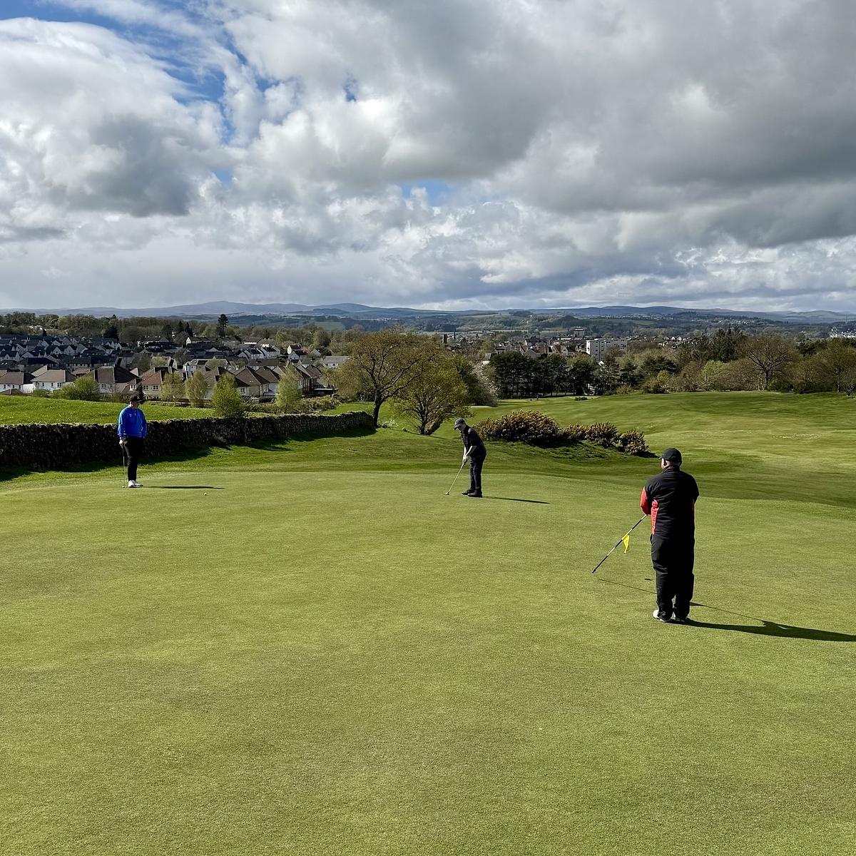 Three people are playing golf on a wide green course under a partly cloudy sky.