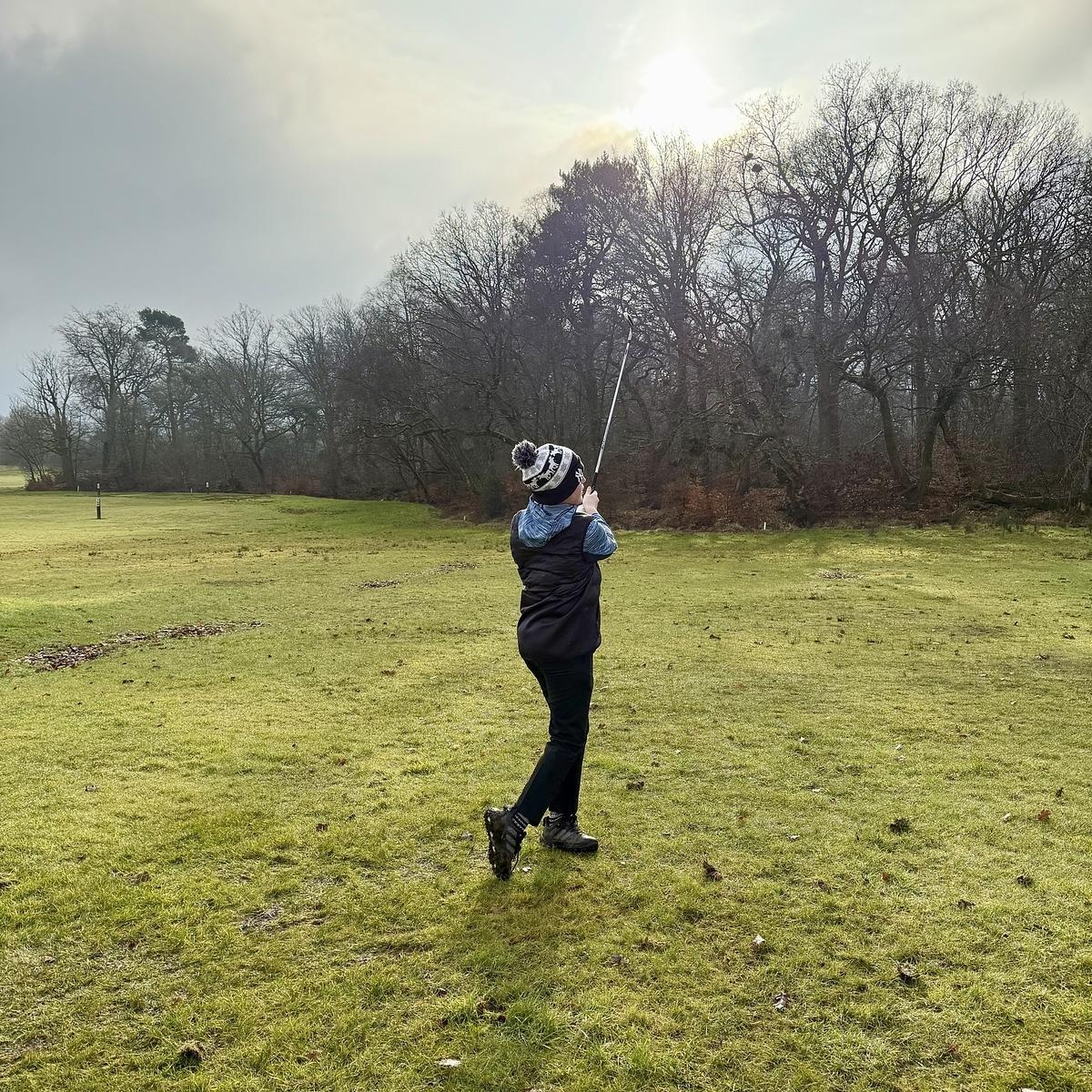 A person is swinging a golf club on a grassy golf course with trees in the background.