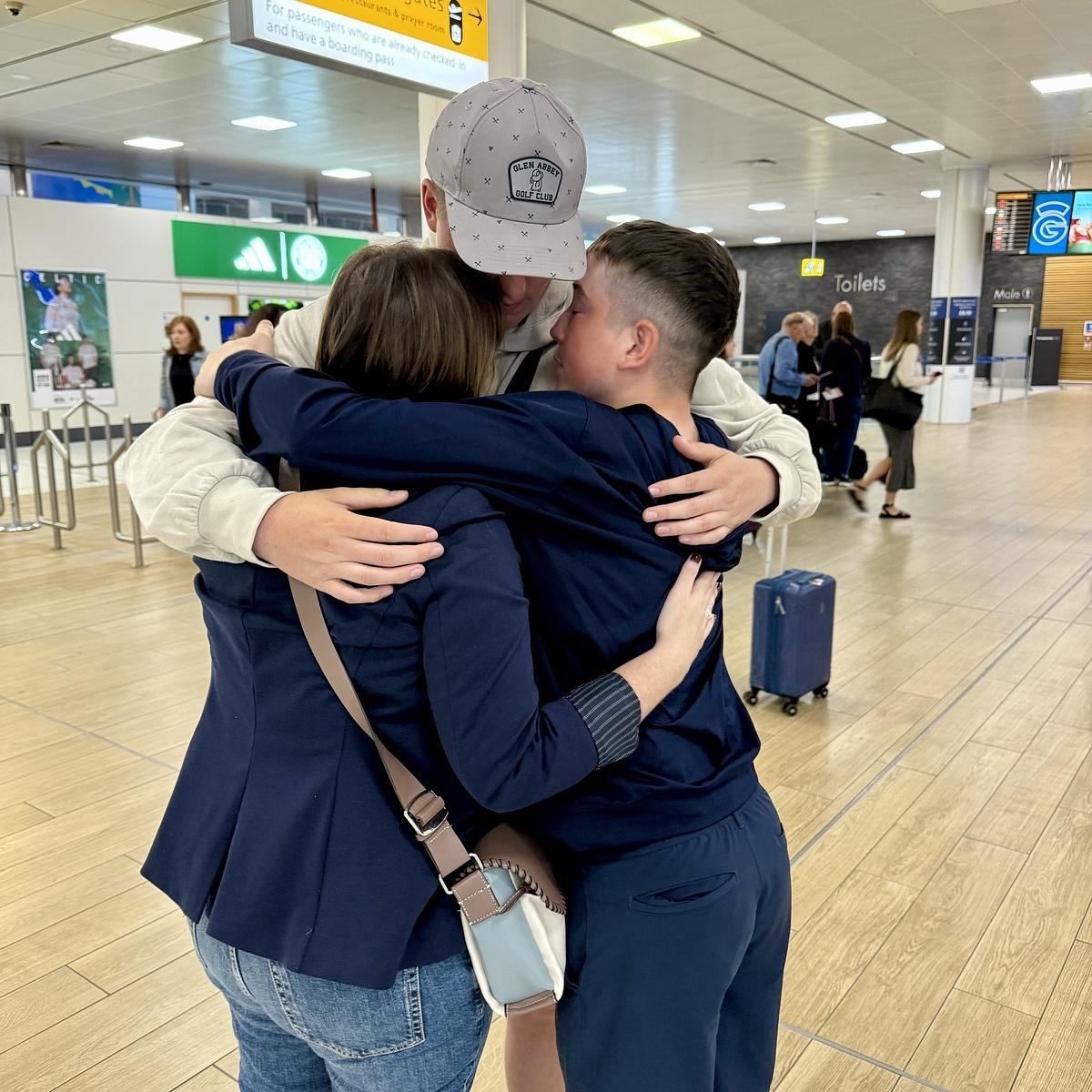 Three people are embracing in a group hug at an airport terminal.