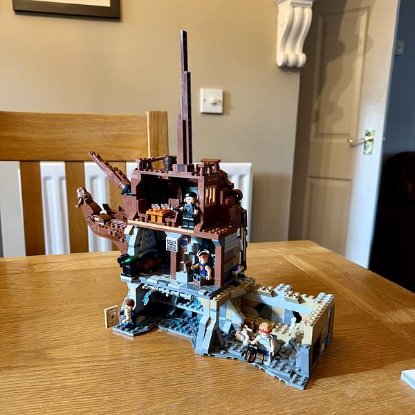 A LEGO pirate shipwreck set displayed on a wooden dining table, featuring a tilted brown ship hull built into gray rocky cliffs with several minifigures positioned throughout the scene, including characters on the upper deck and in a cave-like area below.