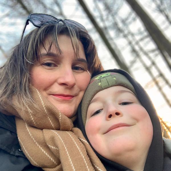 A woman and a child, both wearing warm clothing, smile while taking a selfie outdoors with trees in the background.