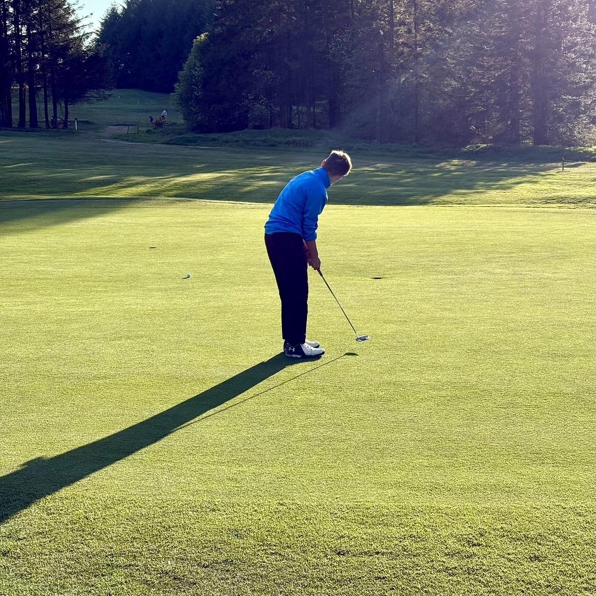 A person is putting on a golf green surrounded by trees under the bright sunlight.