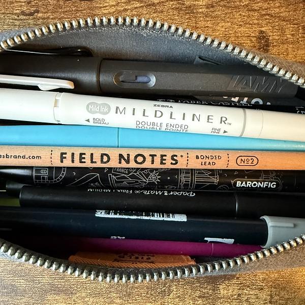 A zippered pouch contains various pens, markers, and a pencil above a Field Notes booklet.