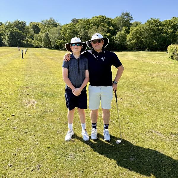Two people wearing wide-brimmed hats and sunglasses stand together on a golf course, with one holding a golf club.