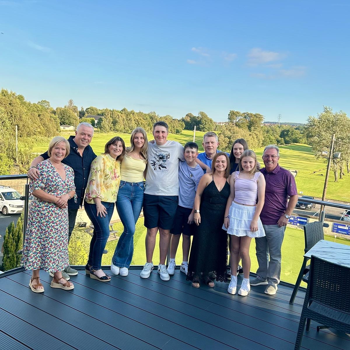 A group of people is smiling and posing together on a deck with a scenic green landscape in the background.
