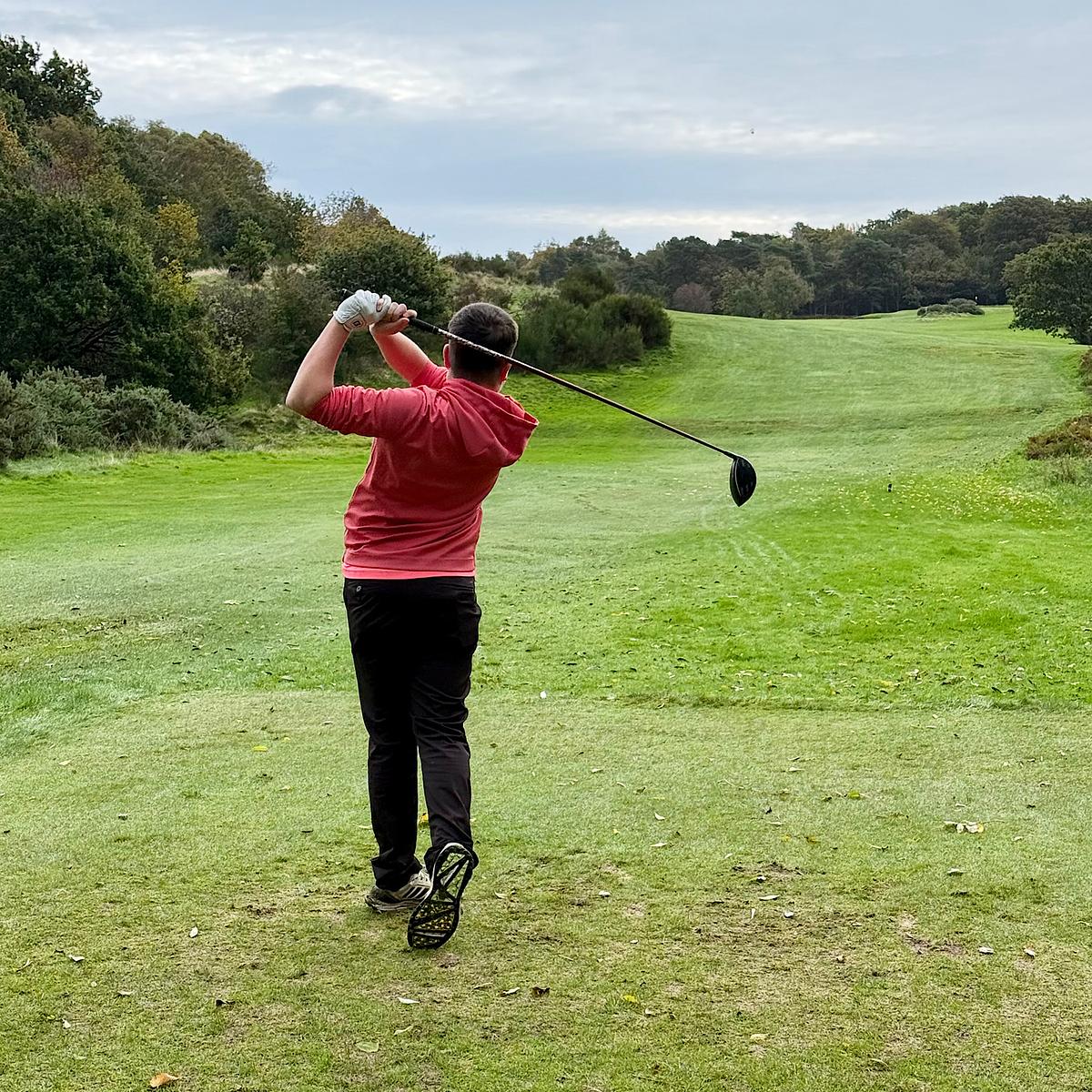 A golfer in a red top and black pants follows through after driving a ball down a lush green fairway lined with trees.