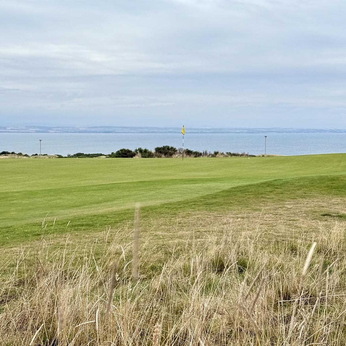 A golf course lies adjacent to a calm ocean under a cloudy sky.