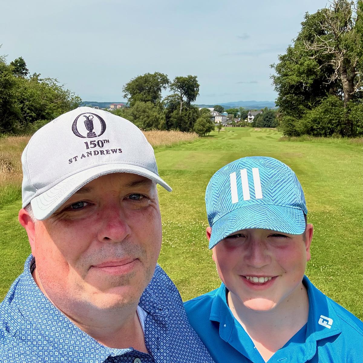 Two people smiling on a golf course, the adult wearing a 150th St Andrews Open Championship cap and the youth in an Adidas cap.