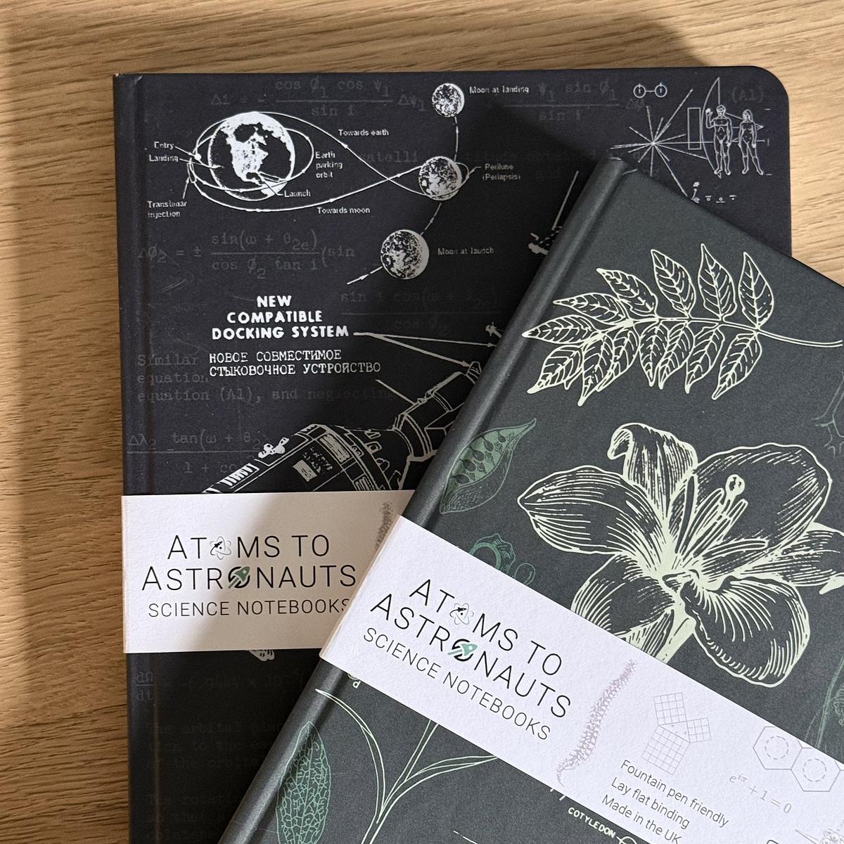 Two science-themed notebooks are displayed on a wooden surface, featuring intricate scientific and botanical designs on their covers.
