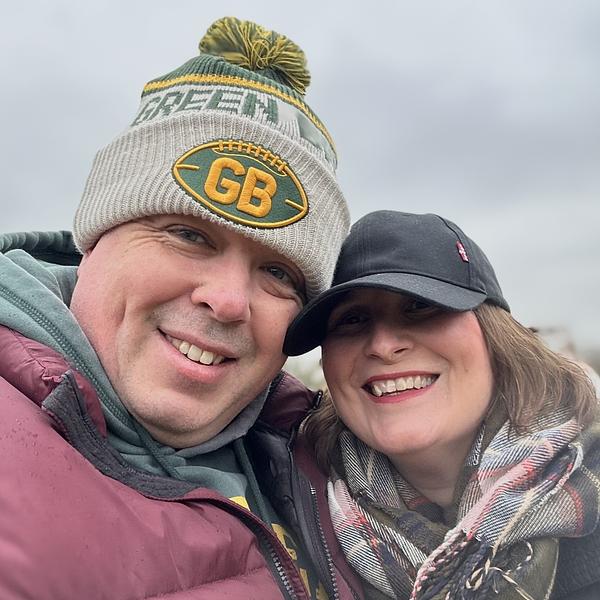 A smiling couple is wearing winter clothing, including a knit hat with a sports logo and a black cap, outdoors.