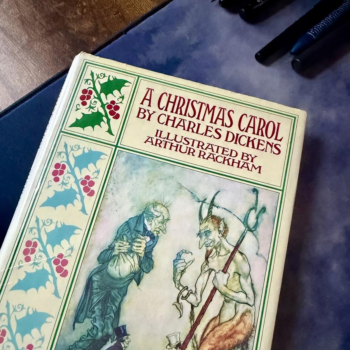 A copy of "A Christmas Carol" by Charles Dickens, illustrated by Arthur Rackham, is lying on a table with pens nearby.