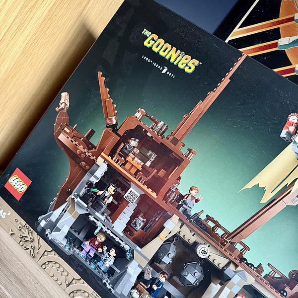 A LEGO Ideas set based on The Goonies, showing the box art featuring One-Eyed Willy's pirate ship built in brown and grey bricks. Multiple minifigures representing characters from the 1985 film are arranged across the multi-level ship interior. The box sits on a wooden surface, with a retro space-themed poster partially visible in the background.
