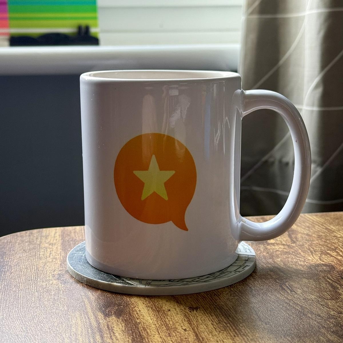 A white mug with an orange speech bubble and green star design rests on a coaster on a wooden table.
